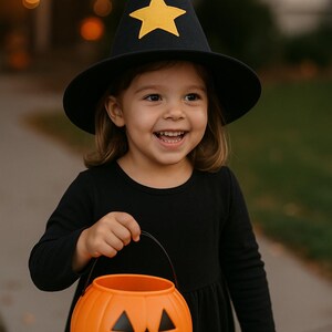 Könnte beinhalten: Ein Kind in einem schwarzen Hexenkostüm, einschließlich eines Hutes mit einem gelben Stern und einem schwarzen Kleid, lächelt, während es einen orangefarbenen Kürbis-Eimer hält. Die Szene spielt sich im Freien ab, wahrscheinlich zu Halloween.