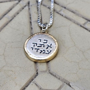 May include: A silver necklace with a circular pendant bearing Hebrew script. The pendant has a gold-coloured rim and a silver centre. The chain is a fine silver link design. The necklace is shown on a textured surface.