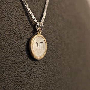 May include: A silver chain necklace with a round pendant featuring the Hebrew letter 'Heh' in a gold circle.