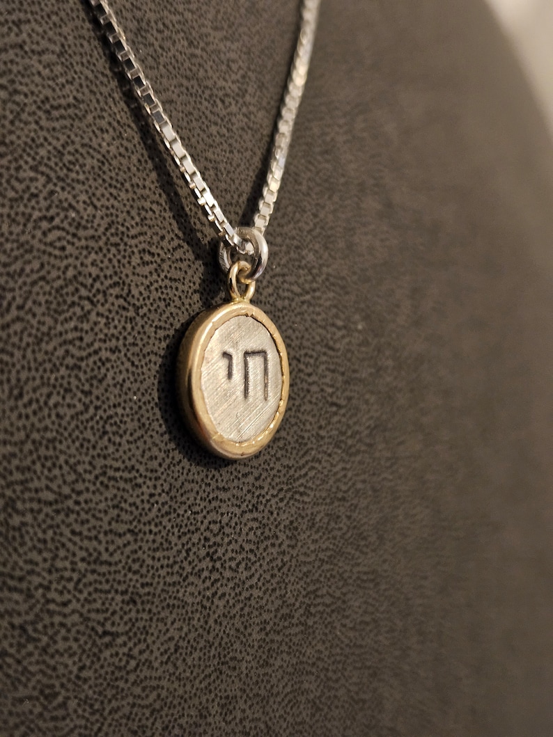 May include: A silver chain necklace with a round pendant featuring the Hebrew letter 'Heh' in a gold circle.