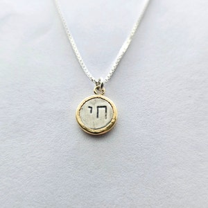 May include: A silver chain necklace with a round pendant featuring the Hebrew letter 'Chai' in black on a silver background. The pendant has a gold rim.