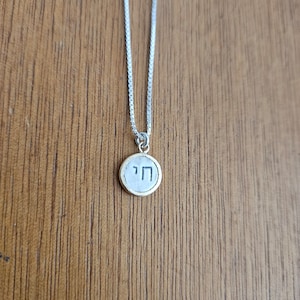 May include: A silver chain necklace with a round pendant featuring the Hebrew letter "Chai" in black lettering on a gold background.