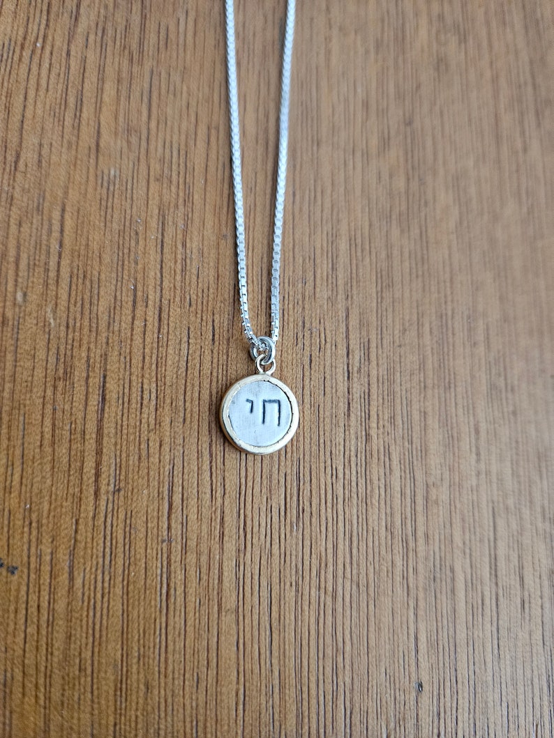 May include: A silver chain necklace with a round pendant featuring the Hebrew letter "Chai" in black lettering on a gold background.