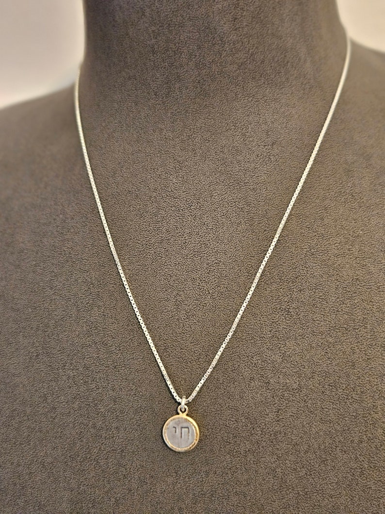 May include: A silver chain necklace with a round pendant featuring the Hebrew letter 'Chai' in silver with a gold border.