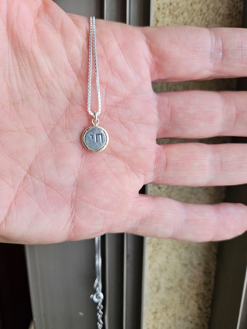 May include: A silver chain necklace with a small round pendant. The pendant is silver with a gold rim and has the Hebrew letter "Chai" inscribed on it.