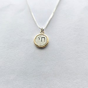 May include: A silver chain necklace with a round pendant featuring the Hebrew letter 'Chai' in black lettering on a silver background with a gold rim.