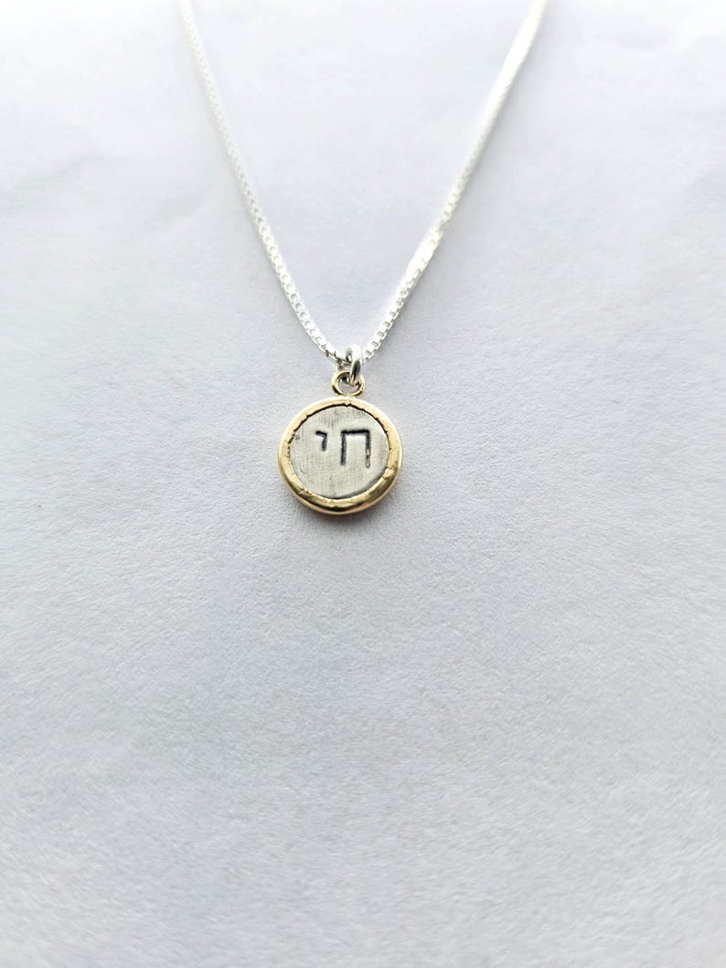 May include: A silver chain necklace with a round pendant featuring the Hebrew letter 'Chai' in black lettering on a silver background with a gold rim.