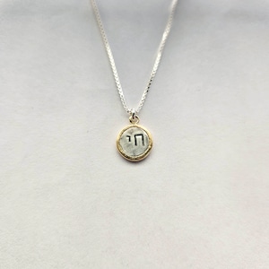 May include: A silver chain necklace with a small, round pendant. The pendant is gold-rimmed and features the Hebrew letter "nun" in silver.