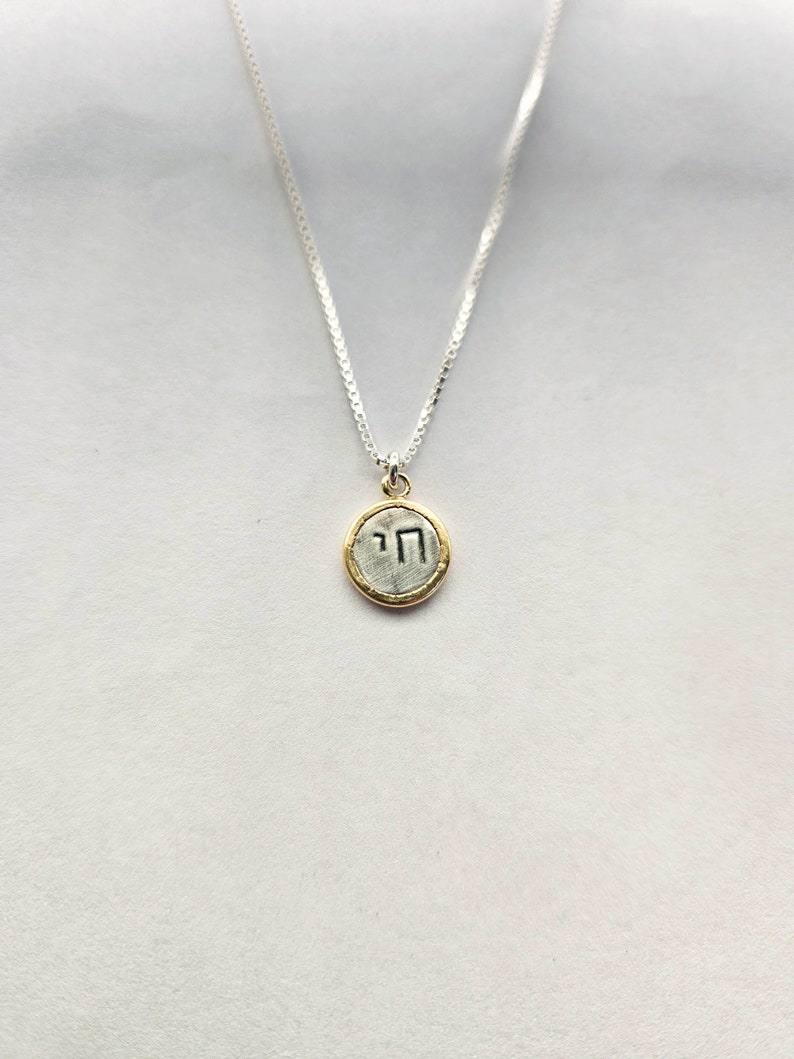 May include: A silver chain necklace with a small, round pendant. The pendant is gold-rimmed and features the Hebrew letter "nun" in silver.