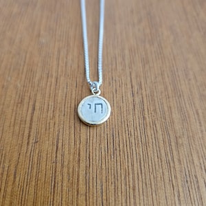 May include: A silver chain necklace with a round pendant featuring the Hebrew letter 'Chai' in black lettering on a silver background. The pendant has a gold rim.