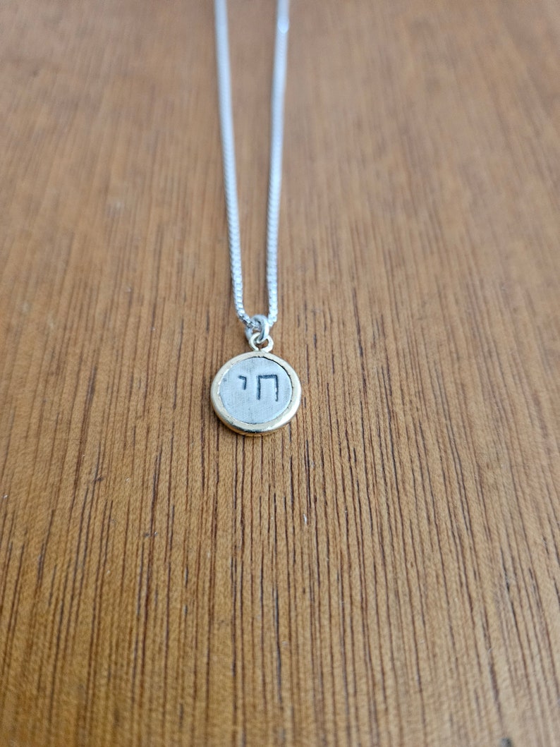May include: A silver chain necklace with a round pendant featuring the Hebrew letter 'Chai' in black lettering on a silver background. The pendant has a gold rim.