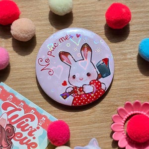 Puede incluir: Un botón redondo rosa claro con un conejo de dibujos animados con un vestido de lunares rojos y un cubo de pintura. El botón tiene el texto "No puc més" y está rodeado de pompones de colores y una flor rosa.