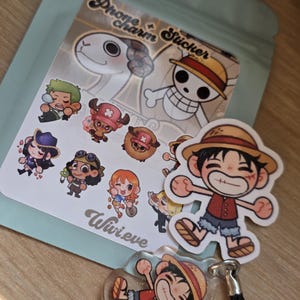 Op de afbeelding: Een verzameling stickers en een telefoonbedel in anime-stijl met personages uit een populaire mangaserie. De stickers tonen verschillende personages met unieke outfits en uitdrukkingen. De telefoonbedel heeft een zwart koord en een heldere, glinsterende afwerking.