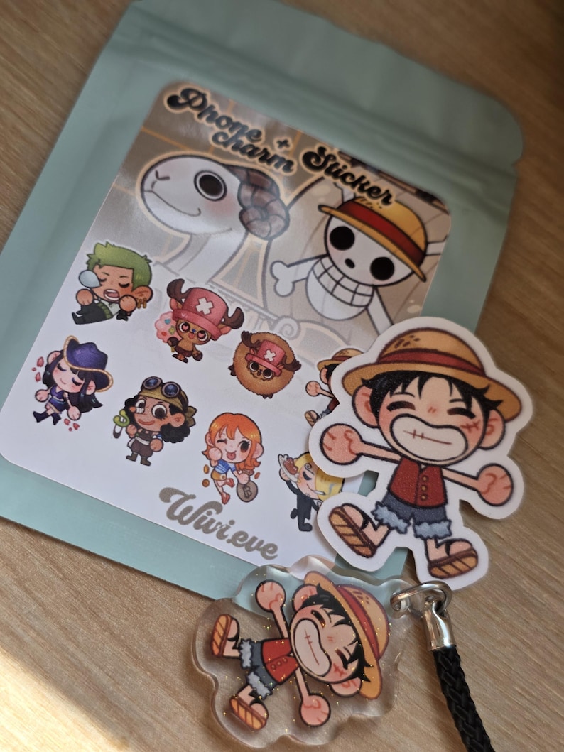 Op de afbeelding: Een verzameling stickers en een telefoonbedel in anime-stijl met personages uit een populaire mangaserie. De stickers tonen verschillende personages met unieke outfits en uitdrukkingen. De telefoonbedel heeft een zwart koord en een heldere, glinsterende afwerking.