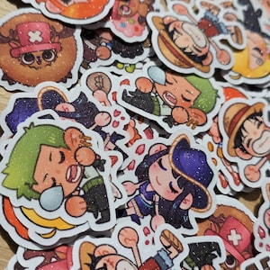 Op de afbeelding: Een verzameling kleurrijke, glinsterende stickers met verschillende stripfiguren. De stickers tonen personages met onderscheidende kenmerken, waaronder strohoeden, zwaarden en medische symbolen. De ontwerpen zijn wit omlijnd, met een glanzende afwerking.