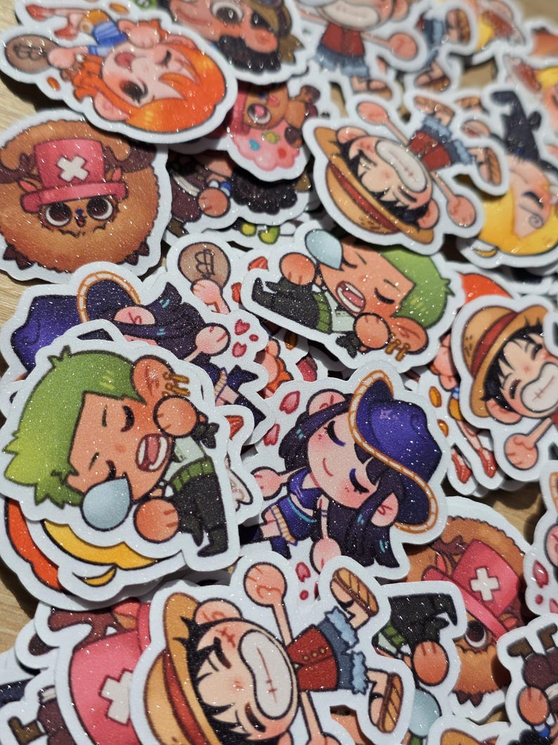 Op de afbeelding: Een verzameling kleurrijke, glinsterende stickers met verschillende stripfiguren. De stickers tonen personages met onderscheidende kenmerken, waaronder strohoeden, zwaarden en medische symbolen. De ontwerpen zijn wit omlijnd, met een glanzende afwerking.