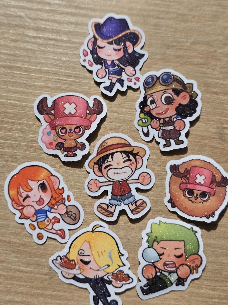 Op de afbeelding: Een verzameling kleurrijke stickers in cartoonstijl met personages uit de anime-serie One Piece. De stickers tonen verschillende personages in dynamische poses, met levendige kleuren en gedetailleerde ontwerpen. De stickers liggen op een lichtbruin oppervlak.