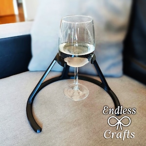 Könnte beinhalten: Ein schwarzer Weinglashalter aus Metall mit einem Glas Weißwein darin. Der Halter ist so konzipiert, dass er das Glas sicher hält und ein Umkippen verhindert. Der Halter steht auf einem hellgrauen Sofa.