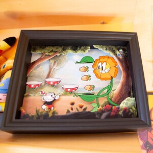 Può includere: Un diorama incorniciato con un personaggio dei cartoni animati, Cuphead, in un ambiente boschivo. Cuphead tiene in mano una tazza da tè e si trova di fronte a un fiore sorridente con un viso. Ci sono quattro tazze da tè che galleggiano nell'aria.