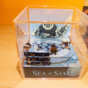 Puede incluir: Una vitrina de acrílico transparente con un diorama en su interior que representa una escena del videojuego Sea of Stars. El diorama presenta personajes pixelados y una gran criatura blanca en forma de nube. La vitrina tiene una base negra con el título del juego, "Sea of Stars", impreso en ella.
