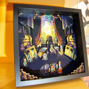 Puede incluir: Un diorama 3D enmarcado que representa una escena pixelada con personajes y una sala del trono. La obra de arte presenta un marco negro, con una escena interior detallada y colorida. El diorama se exhibe en una estantería de madera, con otras obras de arte visibles en el fondo.