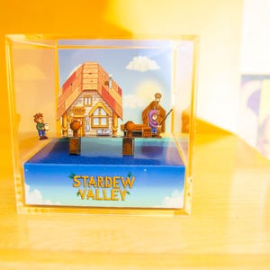 Könnte beinhalten: Eine transparente Acryl-Vitrine mit einem Miniatur-Diorama eines Hauses und einer Figur aus dem Videospiel Stardew Valley. Die Basis des Dioramas ist blau mit dem Text "Stardew Valley" in weiß.