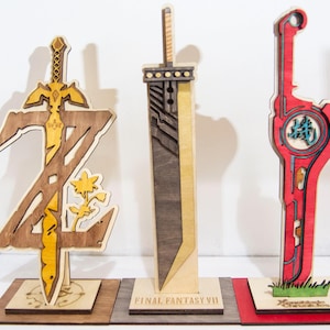 Puede incluir: Tres espadas decorativas de madera. La primera es dorada y marrón con un diseño estilizado de "Z". La segunda es una espada gris, marrón y beige con "FINAL FANTASY VII" en la base. La tercera es roja con texto japonés.