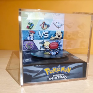 Può includere: Una teca in acrilico trasparente con una base rotante che presenta una cartuccia di gioco Pokemon Edizione Platino. La teca è decorata con un'immagine stampata di personaggi Pokemon e il testo "Pokemon Edicion Platino".