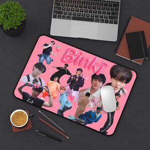 May include: Pink rectangular mousepad featuring a collage of images of people and the word "Bink" in pink bubble letters. A computer mouse is on the right side of the mousepad. A cup of coffee and a laptop are also visible.