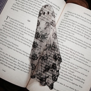 Puede incluir: Un marcap&aacute;ginas con forma de fantasma en blanco y negro hecho de encaje, colocado en un libro abierto. El marcap&aacute;ginas tiene una silueta de fantasma con recortes para los ojos y la boca. Las p&aacute;ginas del libro muestran texto. La palabra "GOTHIKANA" es visible.