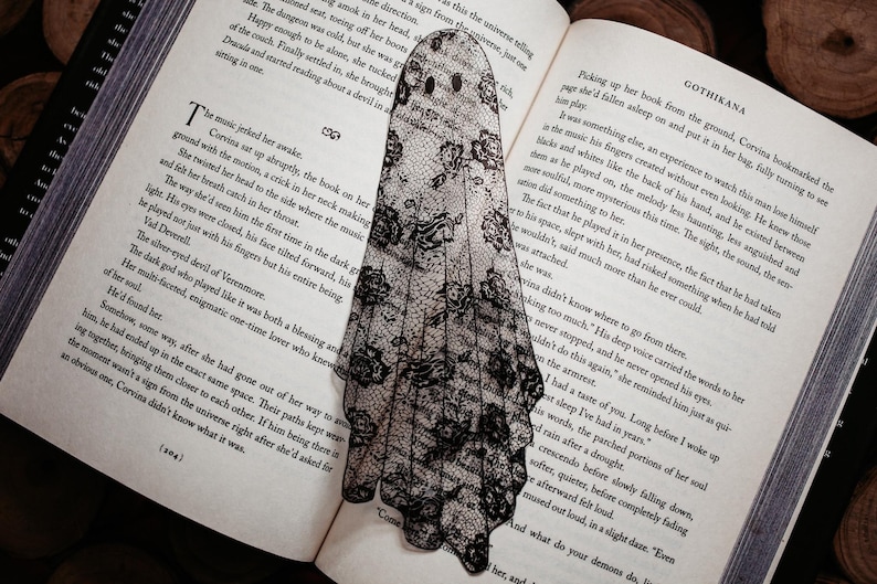 Puede incluir: Un marcap&aacute;ginas con forma de fantasma en blanco y negro hecho de encaje, colocado en un libro abierto. El marcap&aacute;ginas tiene una silueta de fantasma con recortes para los ojos y la boca. Las p&aacute;ginas del libro muestran texto. La palabra "GOTHIKANA" es visible.