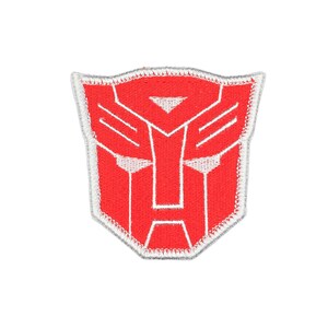 May include: A shield-shaped embroidered patch featuring the red Autobot insignia from the Transformers franchise. The design consists of a stylised robotic face mask outlined in white thread against a solid red background with a white border.