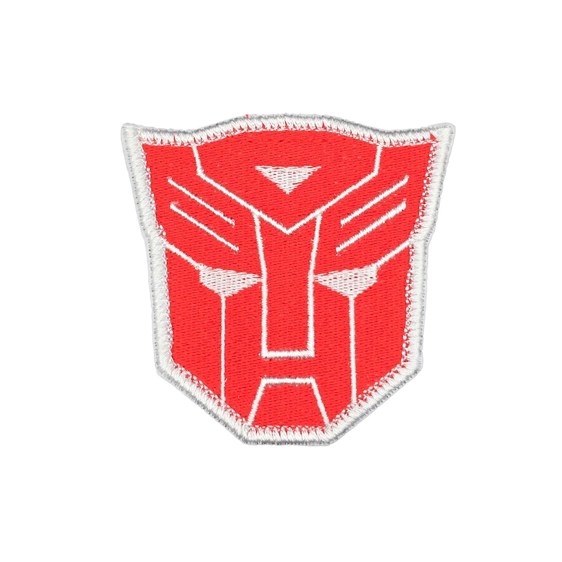 May include: A shield-shaped embroidered patch featuring the red Autobot insignia from the Transformers franchise. The design consists of a stylised robotic face mask outlined in white thread against a solid red background with a white border.