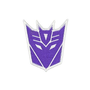 May include: A purple embroidered patch featuring the Decepticon logo from Transformers. The design is shield-shaped with sharp, angular lines and a white border, showcasing a stylised robotic face with pointed ears and a triangular forehead detail.