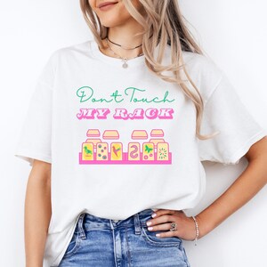 Op de afbeelding: Wit T-shirt met de tekst "Don't Touch My Rack" in roze en groene letters. De tekst wordt vergezeld door een afbeelding van zes kleurrijke potten met verschillende ontwerpen.