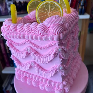 May include: A square-shaped box decorated with pink frosting in a piped cake style, featuring intricate scalloped borders, swags, and small white pearl accents. The top edge is adorned with four yellow lemon slice decorations.