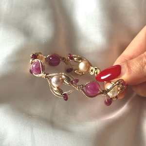 May include: A gold bracelet with a delicate design, adorned with pink and purple gemstones and white pearls. The bracelet is displayed against a white satin backdrop, highlighting its intricate details and colour scheme.