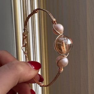 May include: A gold-coloured wire bracelet with a clear, round bead with internal inclusions, and two smaller, pinkish-white beads. The bracelet is held by a hand with red nail polish, highlighting the detailed wirework and bead arrangement.