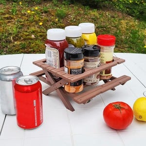 May include: A miniature brown picnic table holds various condiment bottles and spice jars. Two aluminum cans and a red can sit to the left. A red tomato and a yellow lemon are on the right. The background is a blurred green lawn.