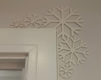 May include: White snowflake decorations of varying sizes are arranged around the corner of a white door frame. The snowflakes are cut out and appear to be made of a lightweight material, creating a winter holiday theme.