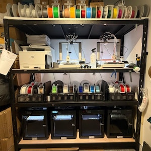May include: A black metal shelving unit holding four 3D printers on the bottom shelf, two open-frame 3D printers and a white printer on the middle shelf, and numerous colourful plastic filament spools organised on the top and middle shelves.
