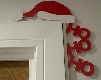 May include: A red and white Santa hat door decoration with the words "HO HO HO" in red. The decoration is placed on the top corner of a white door frame, creating a festive holiday display.