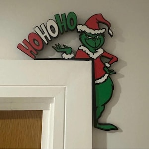 May include: A Christmas door decoration featuring the Grinch in a red and white Santa hat and jacket. The words "HO HO HO" are above the Grinch's head in red and green.