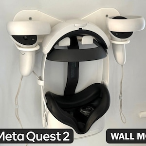 Puede incluir: Un soporte de pared blanco que sostiene un casco de realidad virtual Meta Quest 2 y dos mandos blancos a juego. El casco cuenta con una interfaz facial y una correa negras, mientras que los mandos se sitúan en los brazos del soporte.