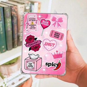 May include: A pink phone case with various stickers, including roses, a crown, and the words "Book Boyfriends," "Smut," and "Spicy." The case has a clear border and is held in a hand.