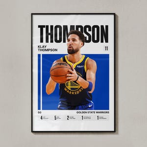 May include: A framed poster of Klay Thompson, a Golden State Warriors basketball player. The image shows Thompson holding a basketball, wearing a blue and gold jersey. The poster includes the name "THOMPSON" in large black letters and various accolades.