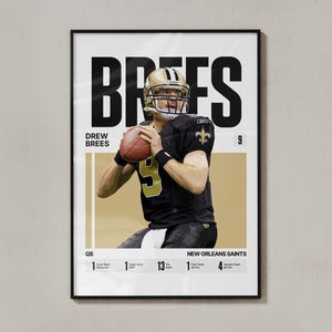 May include: A framed poster of Drew Brees in a New Orleans Saints uniform, holding a football. The poster includes the name "BREES" in large black letters, along with statistics and the team name. The colour scheme is black, gold, and white.