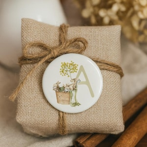 May include: A small, square package wrapped in hessian and tied with twine. A round, white button with a watercolour illustration of bunnies in a basket and the letter "A" is affixed. Cinnamon sticks are in the foreground.