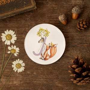 Pode incluir: Um prato de cerâmica branco com uma ilustração em aquarela de uma raposa, uma borboleta e elementos florais. A letra "A" está incorporada no design. O prato tem um tema de floresta e é rodeado por margaridas, bolotas e uma pinha.