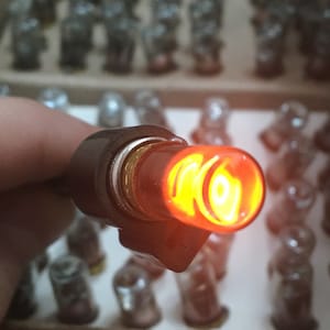 May include: A close-up of a lit red light bulb with a black metal base. The bulb is surrounded by many other unlit bulbs in a box.